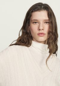 Maglione in cotone chiaro lavorato a trecce con scollo a V profondo e colletto a imbuto a strati. La texture morbida e la vestibilità rilassata aumentano il comfort e il calore.