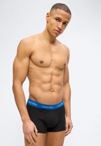 Μυώδης άνδρας με κοντά μαλλιά που φοράει μαύρα εσώρουχα τύπου boxer με μπλε μέση Calvin Klein, στέκεται σε απλό φόντο.