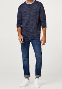 Maglione lungo a maniche con texture in blu navy e colletto grigio, abbinato a jeans blu con risvolto e sneakers bianche.