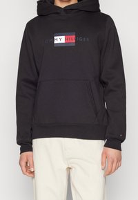 Black hoodie with a front pocket and drawstring hood. Features a logo in red, white, and blue on the chest. Soft fabric texture.