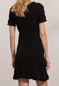 Abito nero a maglia con maniche corte a volant, con dettagli nella parte superiore strutturata e orlo a balze svasato. Il design è aderente e arriva fino al ginocchio.