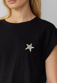 Czarny t-shirt z krótkim rękawem, ozdobiony białą łatką w kształcie gwiazdki morskiej z misternymi niebieskimi i zielonymi detalami na przedniej części.