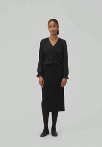 Blusa preta com mangas longas em balão e botão na frente, emparelhada com uma saia lápis preta ajustada, usada com meias pretas e sapatos.