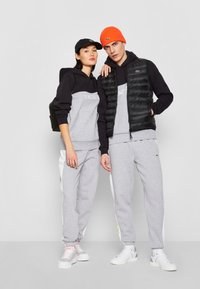 Grijze joggingbroeken met witte zijkanten, bijpassende grijze hoodies met zwarte accenten, een zwarte gewatteerde vest en een feloranje beanie; accessoires zijn onder andere een zwarte rugzak.