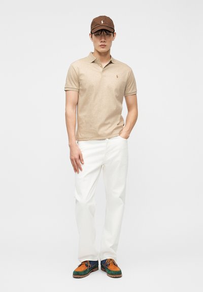 Άνδρας με μπεζ πόλο πουκάμισο, άσπρο παντελόνι, πολύχρωμα παπούτσια και καφέ καπέλο του μπέιζμπολ, στέκεται με το ένα χέρι στην τσέπη.