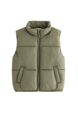 Dreng stående med hænder i lommerne iført olivengrøn polstret vest, cremefarvet kabelstrikket sweater, løse blå jeans og hvide sneakers mod hvid baggrund.