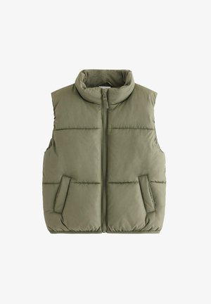 Dreng stående med hænder i lommerne iført olivengrøn polstret vest, cremefarvet kabelstrikket sweater, løse blå jeans og hvide sneakers mod hvid baggrund.