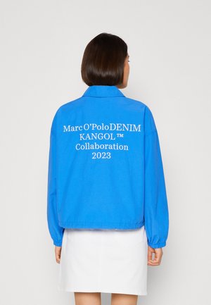 Personne aux cheveux courts et foncés portant une veste bleu vif avec l'inscription « Marc O’Polo DENIM KANGOL™ Collaboration 2023 » imprimée au dos, associée à une jupe blanche.