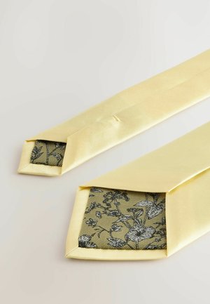 Gul satinfluga med spetsig ände som avslöjar en svart blommönstrad foder på en texturerad bakgrund. Slät yta, elegant design.