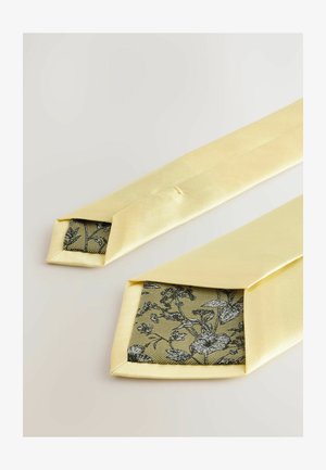 Gul satinfluga med spetsig ände som avslöjar en svart blommönstrad foder på en texturerad bakgrund. Slät yta, elegant design.