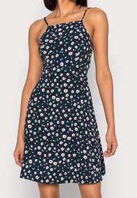 Žena v námořnické modré bezramínkové šaty s tenkými ramínky a bílým květinovým potiskem sedmikrásek, stojící s uvolněnýma rukama podél těla.