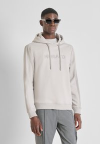 Sudadera gris clara con capucha y logo bordado "MORATO" en el pecho. Cuenta con puños y dobladillo acanalados, combinada con pantalones grises y gafas de sol.