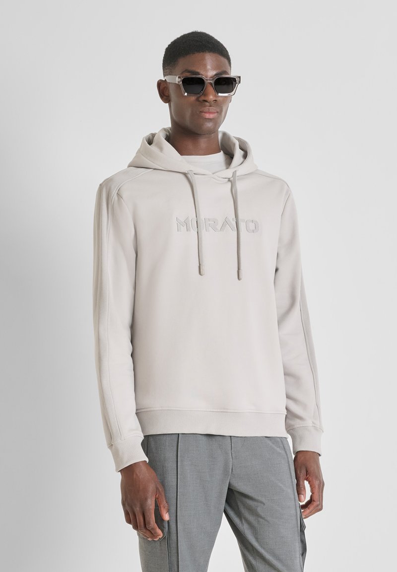 Sudadera gris clara con capucha y logo bordado "MORATO" en el pecho. Cuenta con puños y dobladillo acanalados, combinada con pantalones grises y gafas de sol.