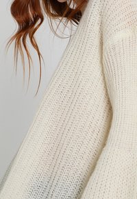 Pull en tricot blanc présentant une coupe ample, un motif texturé et des manches larges. Le tissu semble doux et léger.