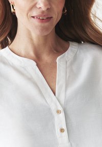 Hvid linnedbluse med V-hals, tre lodrette knapper i naturtræ og en afslappet pasform med subtil tekstur.