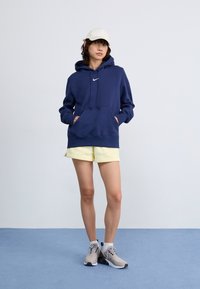 Marineblå hettegenser med snor og frontlomme, kombinert med gule shorts og grå joggesko. Enkelt design med logoaksenter.