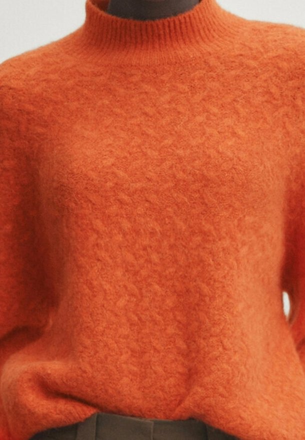 Pull orange à texture douce avec un col rond haut, porté sur un pantalon foncé, montrant le haut du torse et le cou de la personne.