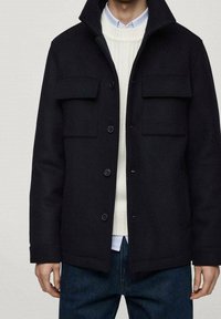 Černá vlněná bunda s dvěma náprsními kapsami, předním zapínáním na knoflíky a vysokým límcem. Nositá přes krémový svetr a modrou košili. Texturovaná tkanina.