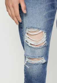 Blå denimjeans med synlige slitasjer og flere revner på knærne, i en lys vask og strukturerte stoff. Hånd hviler i nærheten.