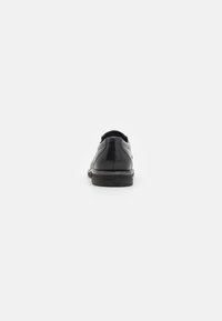 Zapato de cuero negro con punta redondeada, costuras visibles y suela de goma plana. La vista trasera muestra una superficie lisa y un ligero tacón.
