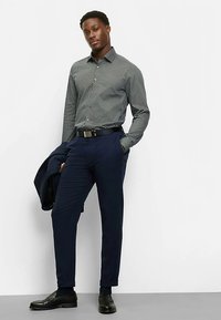 Button-up shirt in dark green with white geometric pattern, paired with navy trousers. Features a black belt and leather shoes.