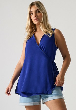 Femme aux longs cheveux blonds portant un haut bleu sans manches noué et un short en jean bleu clair retroussé sur un fond uni.