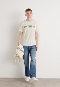 Beige short-sleeve T-shirt with "GANT NH. CT." printed in dark green, paired with light blue jeans. Holding a cream hoodie with similar text.