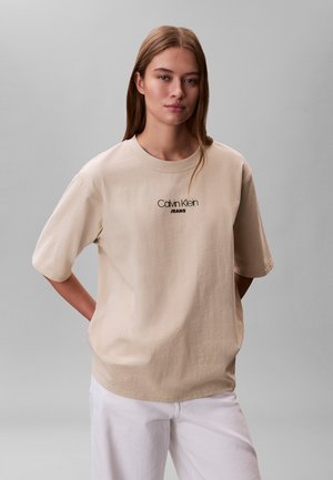 Ung kvinde med lige brunt hår iført beige Calvin Klein Jeans t-shirt og hvide bukser, står med hænderne bag ryggen.