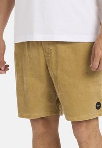 Corduroy shorts in een gouden bruine kleur met een geribbelde textuur. Voorzien van zijzakken en een zwarte, ronde logopatch op de onderzoom.