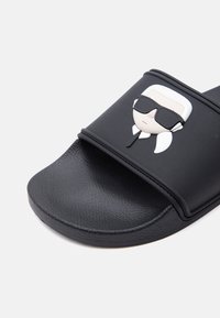 Sandale à glissière noire avec semelle texturée, dotée d'une bride ornée d'un visage stylisé portant des lunettes de soleil et une queue de cheval blanche.