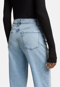 Lichtblauwe denim jeans met een hoge taille, met twee achterzakken en een merklabel. Het model draagt een zwart top met lange mouwen.
