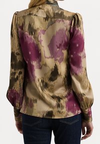 Hedvábná blůza s abstraktním vzorem v purpurové, béžové a hnědé barvě, dlouhými širokými rukávy a úzkými manžetami, nošena s tmavě modrými džíny.