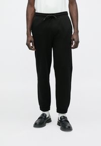 Fekete, elasztikus szegélyű és zsinóros derékú jogger nadrágot viselő személy, fehér zokniban és fekete, könnyen felhúzható cipőben, fehér háttér előtt áll.