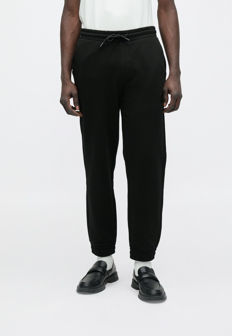 Fekete, elasztikus szegélyű és zsinóros derékú jogger nadrágot viselő személy, fehér zokniban és fekete, könnyen felhúzható cipőben, fehér háttér előtt áll.