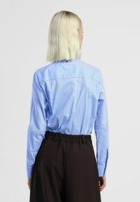 Camicia azzurro chiaro a maniche lunghe con texture liscia e dettaglio sul dietro, abbinata a pantaloni marrone scuro con vita elasticizzata.