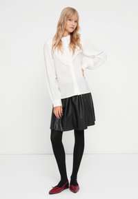 Chemisier blanc à manches longues avec bord en dentelle, fermeture à boutons, associé à une jupe noire en simili cuir, des collants noirs et des ballerines rouges.