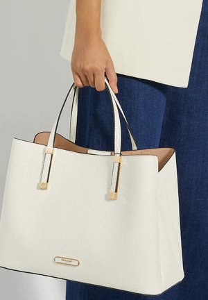 Witte getextureerde handtas met gouden hardware accenten, dubbele bovenhandvatten en een logo plaat aan de voorkant, vastgehouden door een persoon die jeans draagt.