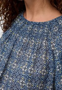 Blouse à motifs bleus avec col smocké. Présente des motifs floraux et géométriques en blanc et jaune sur un fond bleu foncé. Tissu en coton.