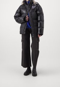 Black puffer jacket with glossy finish, zip closure, and high collar. Paired with dark cargo pants featuring large pockets and contrasting stitching.