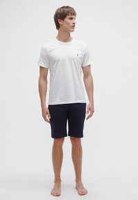 T-shirt branca de algodão com decote redondo, mangas curtas, combinada com calções azul-marinho. O conjunto apresenta um pequeno logótipo no peito.