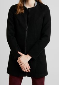 Vrouw in een zwarte ritsjack over een grijze bovenkant, met de handen voor zich gekruist en bordeauxrode broek zichtbaar aan de onderkant.