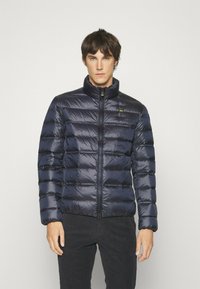 Giacca a vento blu navy con motivo a cuciture verticali, colletto alto, zip frontale e un piccolo logo sul petto. Texture in nylon liscio.