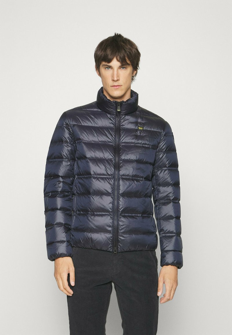 Giacca a vento blu navy con motivo a cuciture verticali, colletto alto, zip frontale e un piccolo logo sul petto. Texture in nylon liscio.