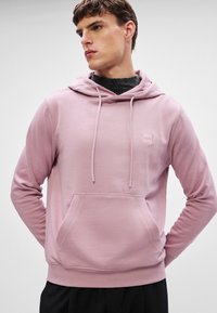 Roosa kapuutsiga särk, millel on känguru tasku ja paelad, silindrilise väikese nurgalogo rinnal. Kangas näeb välja pehme ja tekstuuriga.