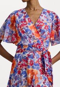 Vestido floral em tecido leve com um padrão vibrante de flores vermelhas, azuis e laranjas, com mangas esvoaçantes e cintura amarrada.