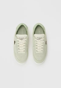 Zapatillas de ante verde claro con cordones blancos, área de los dedos perforada y logotipo de Lacoste en la lengüeta y los laterales, planas sobre fondo blanco.