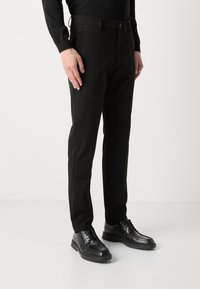 Un pantalon noir à coupe ajustée, fabriqué en tissu lisse. Il dispose d'une taille à boutons, de poches étroites, et est associé à des chaussures noires à lacets.