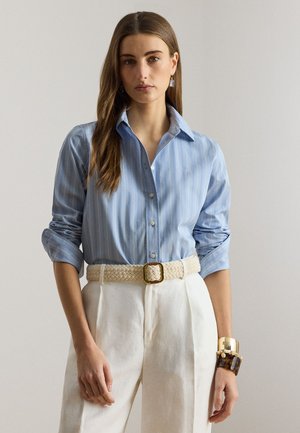 Ženska v modri srajci z belimi črtami z zavihanimi rokavi, belih hlačah z visokim pasom, tkanem pasu in velikih zapestnicah na desnem zapestju.