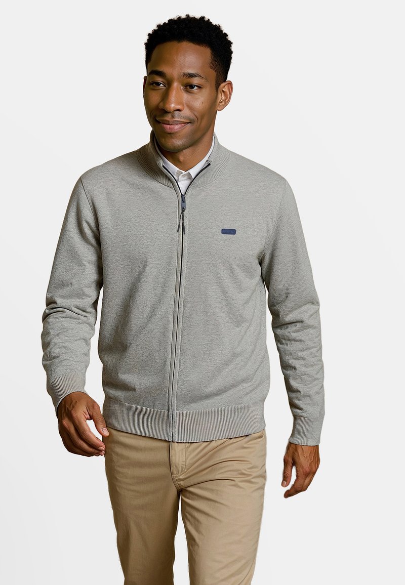 Cardigan zippé gris clair, avec des poignets et un bas côtelés, en tissu lisse, arborant un petit logo bleu sur la poitrine, associé à un pantalon kaki.