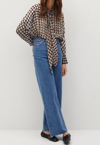 Blusa fluida de estampado marrón y beige con detalle de lazo, combinada con unos vaqueros de mezclilla azul de tiro alto y corte suelto, y unos zapatos planos negros.